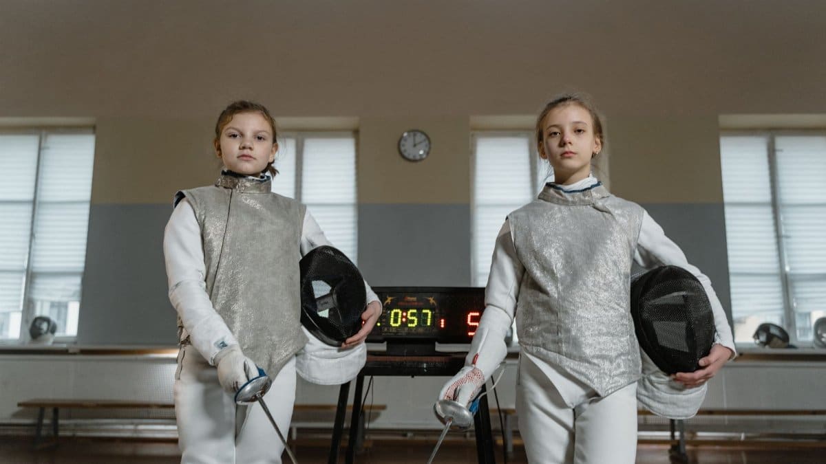 Two young female fencers standing confidently in an indoor arena with fencing gear.