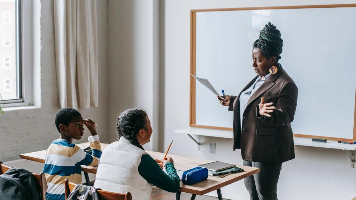 Adult African American female teacher in formal suit standing near whiteboard and explaining new material to multiethnic elementary pupils in light classroom