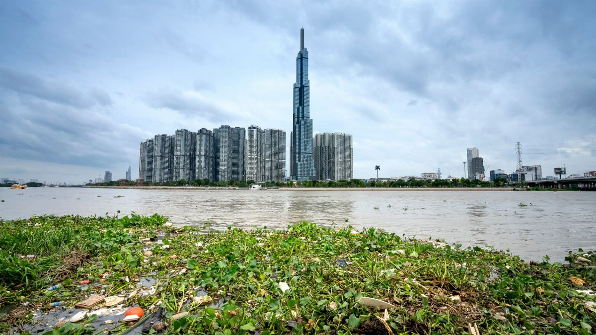 Grassy coast covered in garbage near water and of Ho Chi Minh City in Vietnam with skyscrapers near Landmark 81 building near trees in summer day under cloudy sky