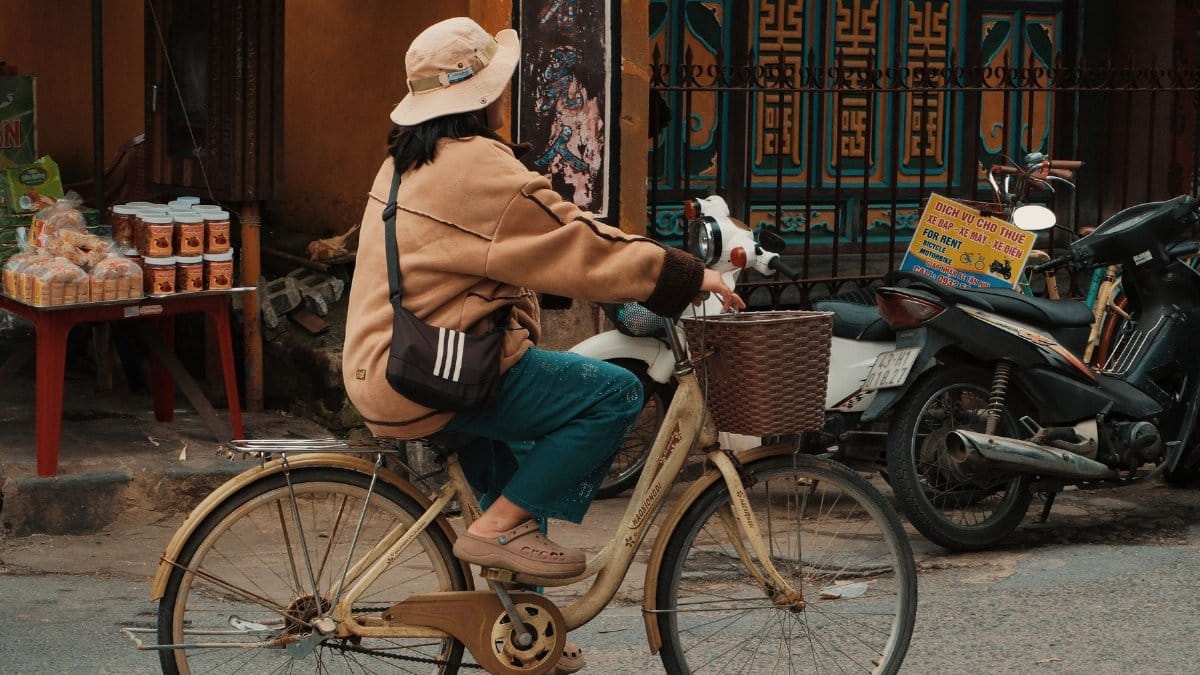 A woman wearing a hat rides a bicycle on a street in Đà Nẵng, Vietnam, featuring a colorful background.