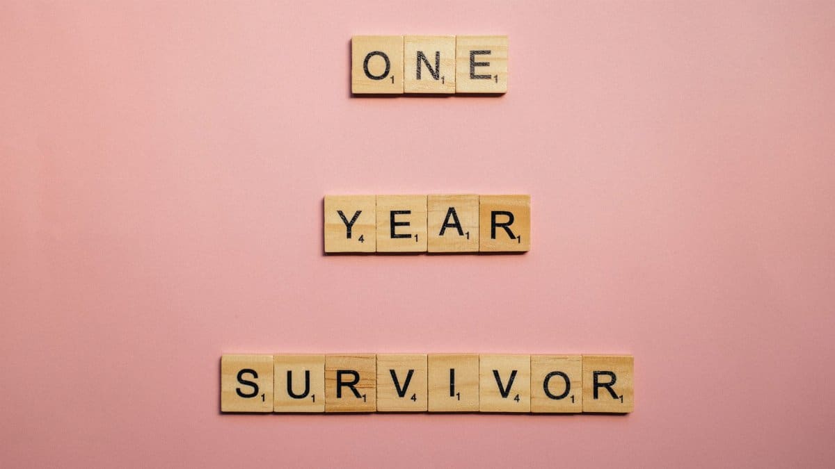 Wooden tiles spell 'One Year Survivor' on a pink background, symbolizing strength and resilience.