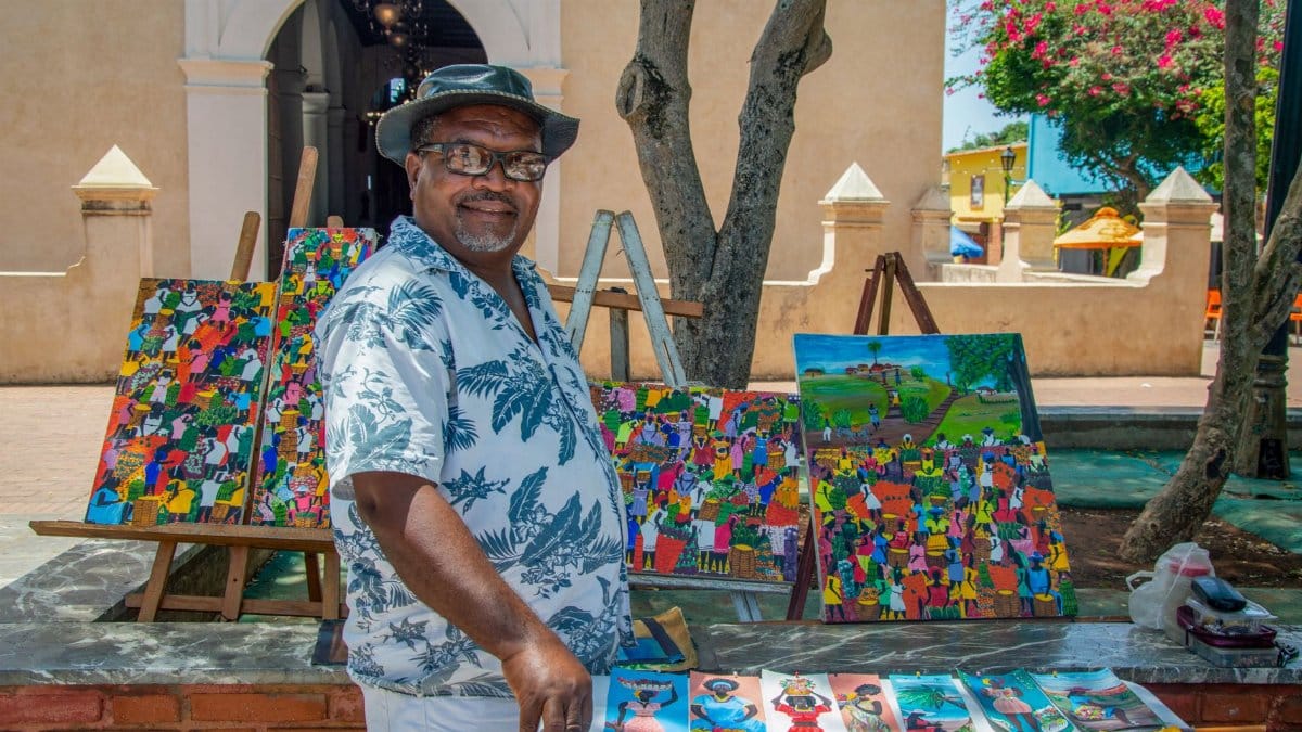 Local artist smiles at his colorful art stall in a sunny outdoor market setting.