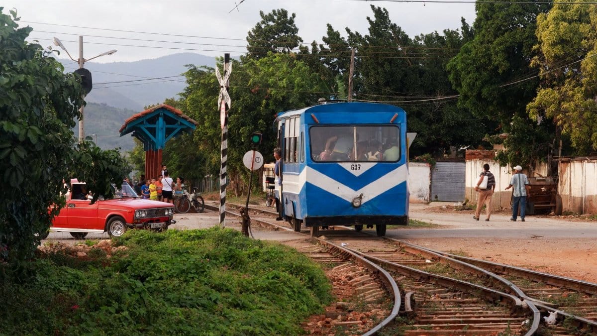 A blue train travels through a rural village with a classic red car and local pedestrians nearby.