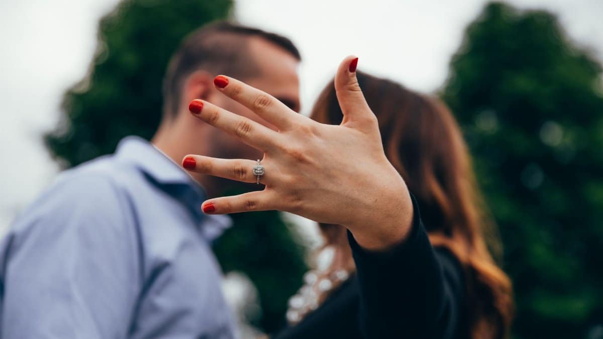 A close-up of a woman's hand showing off an engagement ring with a blurred couple in the background.