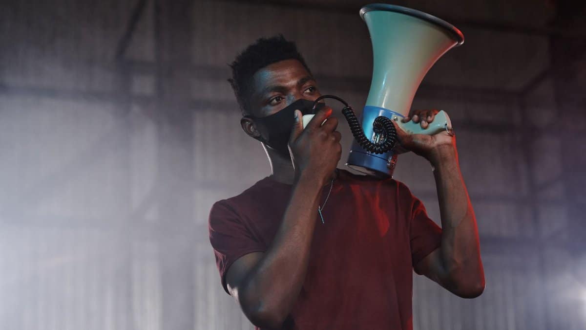 Young man in a red shirt with a facemask using a megaphone indoors.