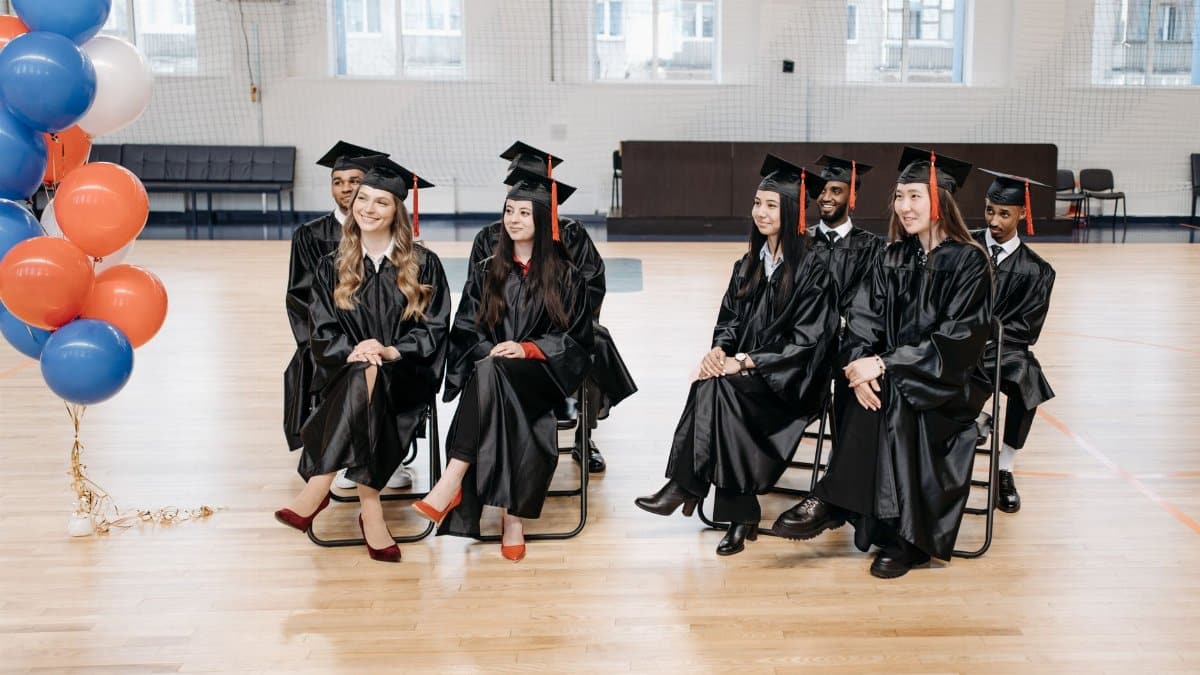 A diverse group of graduates in gowns and caps seated together indoors, celebrating their achievements.
