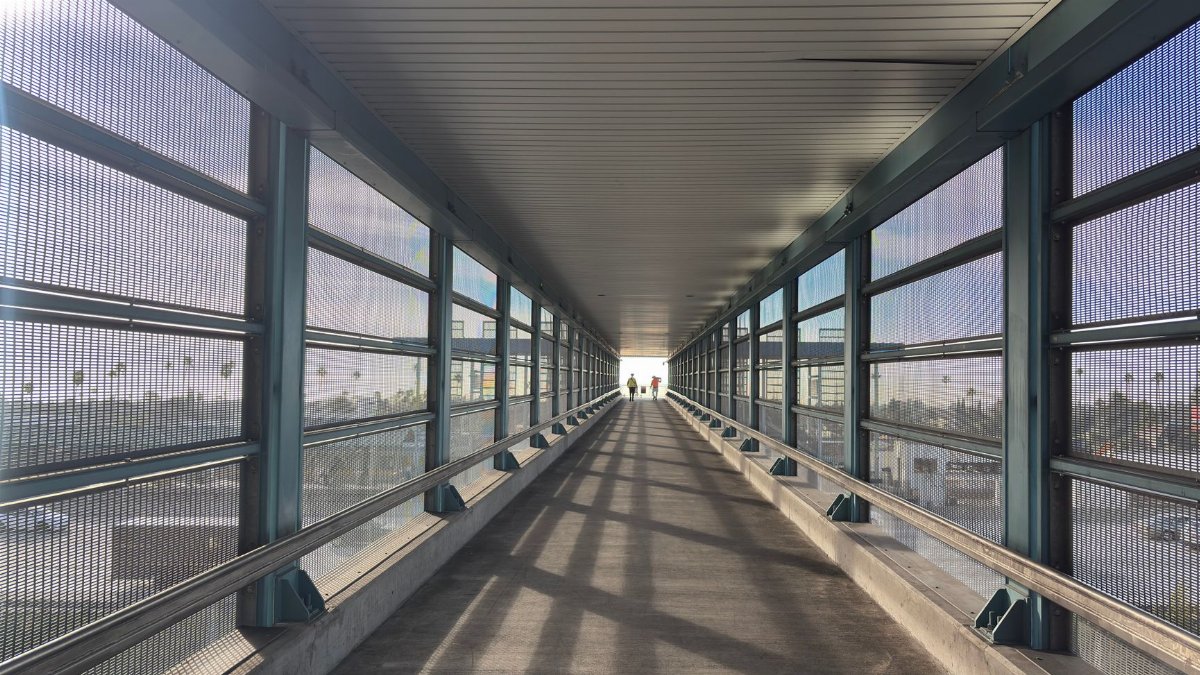 A long pedestrian skywalk with glass walls in a city, leading to a bright exit and featuring two figures walking towards the light.