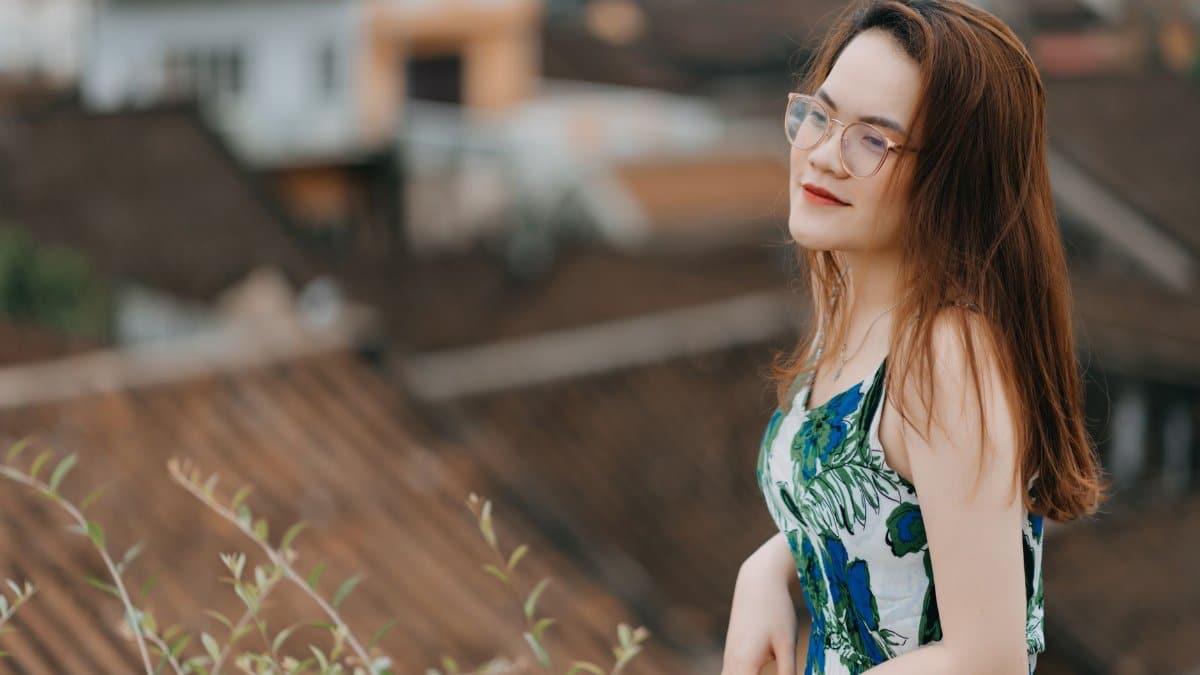 Side view of dreamy young Asian female traveler in stylish dress and eyeglasses standing on viewpoint and admiring old town during sightseeing trip