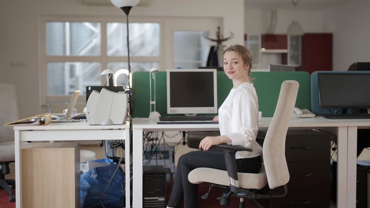 Side view of glad female worker in classy outfit sitting at table on office chair during work in modern workplace and looking at camera