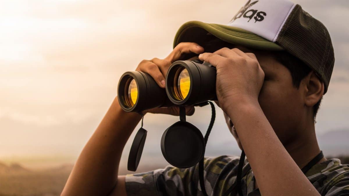 A young man wearing a cap uses binoculars to observe the scenic landscape outdoors during daylight.