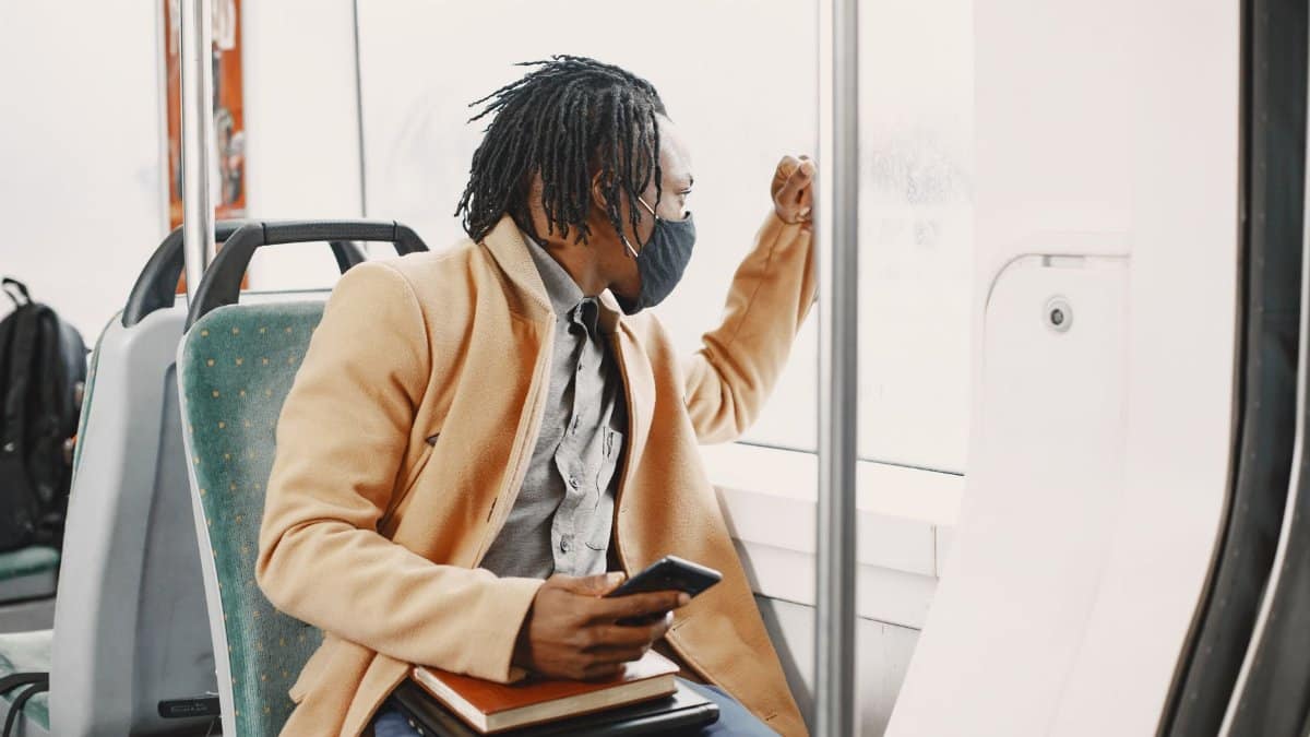 A man in a face mask sits in a bus, looking outside while holding a smartphone.