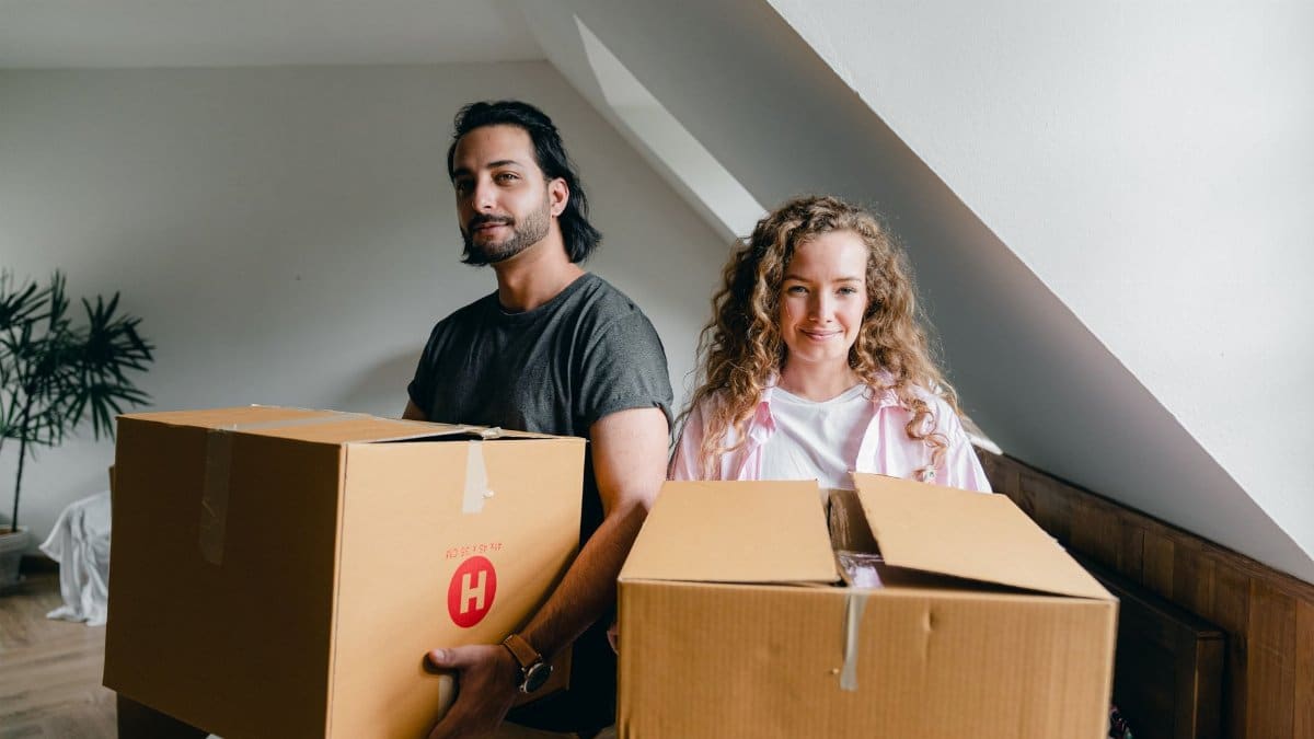 Young woman and ethnic man in casual clothes smiling at camera and holding big boxes during relocation to new apartment