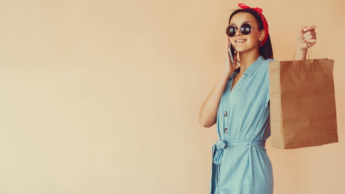 Side view of cheerful stylish young lady in trendy denim dress and stylish red hair band showing paper shopping bag while talking on cellphone in studio on beige background