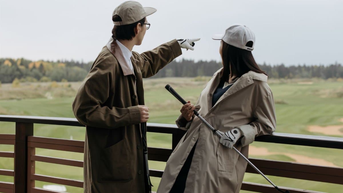 A couple on a balcony overlooking a golf course, wearing coats and caps, and holding a golf club.