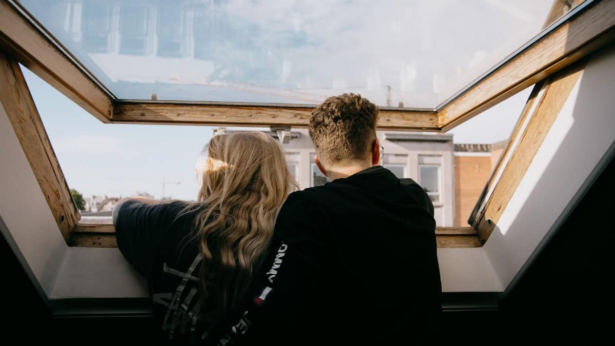 Back view of faceless couple in casual clothes hugging while standing near open window and looking at city