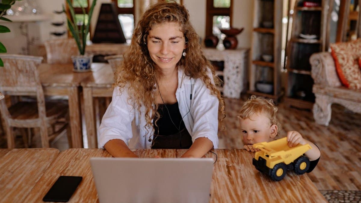 A woman working on a laptop at home with her child playing beside her, creating a balanced family lifestyle.