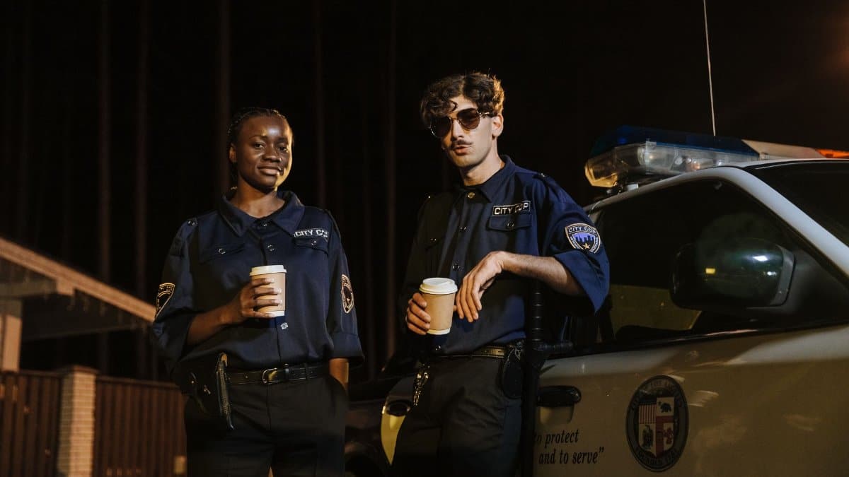 Two police officers standing by a patrol car at night, holding coffee cups and smiling.