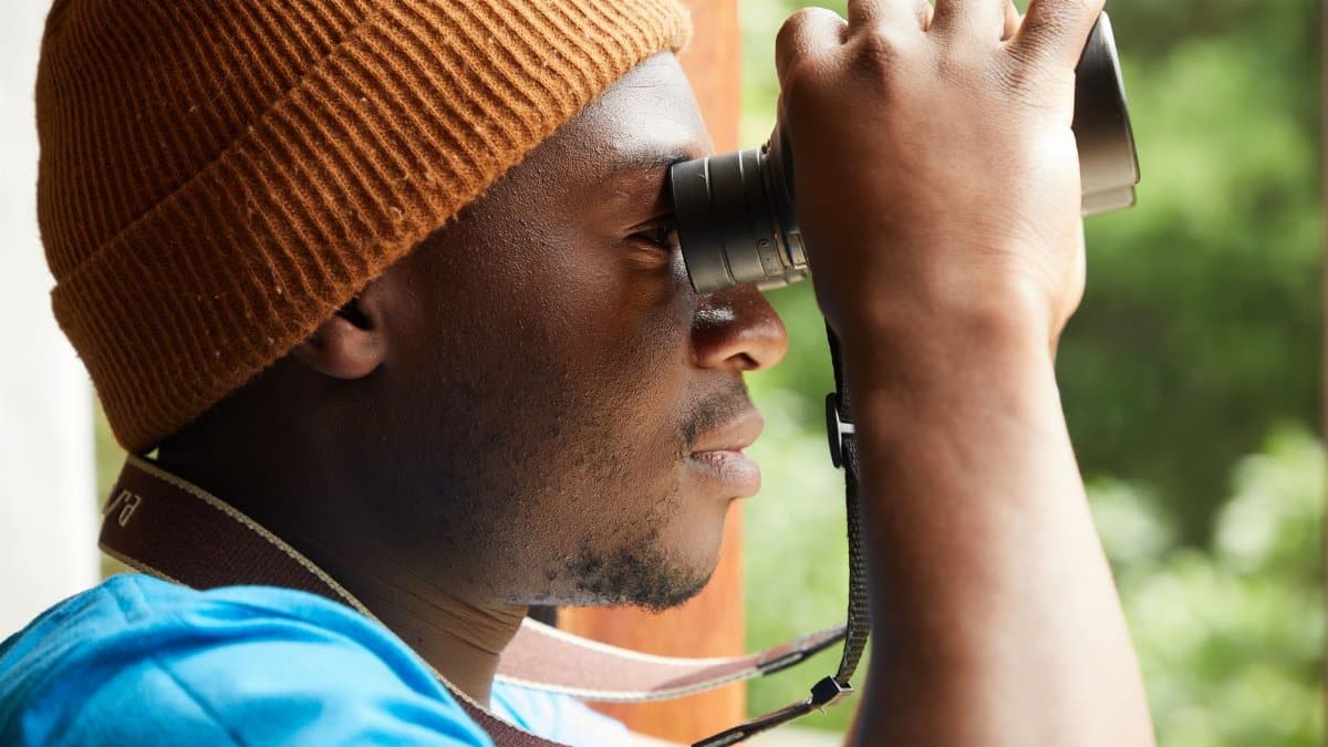 Side view of anonymous African American male in trendy hat looking through binoculars while standing against opened window