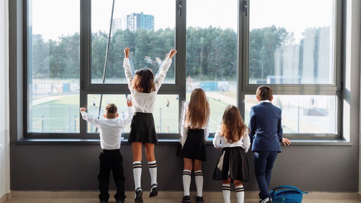 A group of school children in uniform stand by a large window, looking outside.