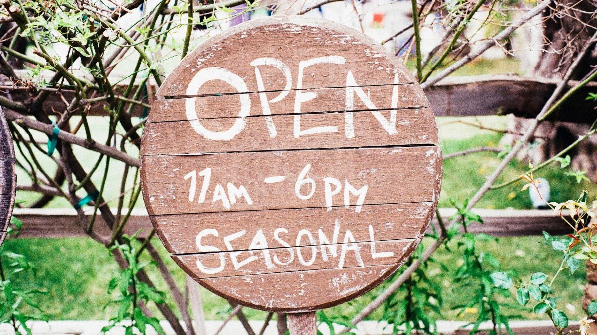 Round wooden sign displaying 'Open 11am-6pm Seasonal' amidst greenery.
