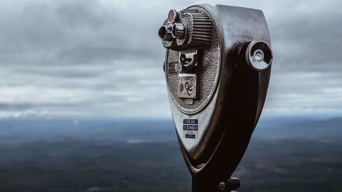 A metal coin-operated binocular telescope on a mountain with a cloudy sky in the background.