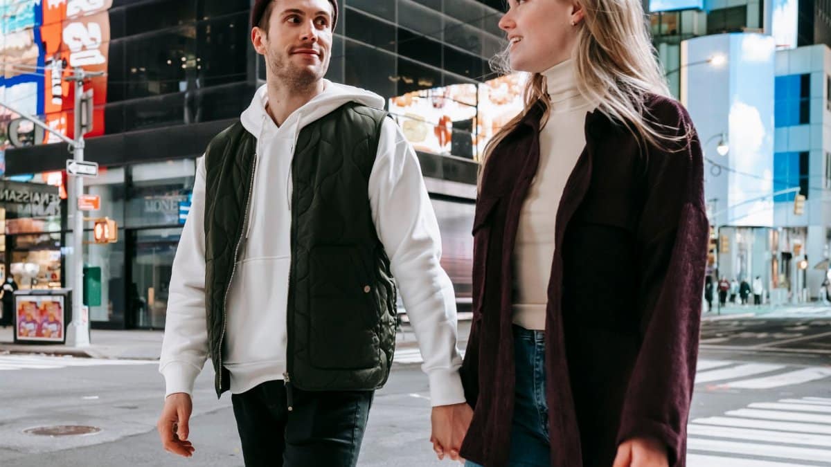 Loving couple holding hands and looking at each other while strolling on pedestrian crossing on street with modern buildings in downtown