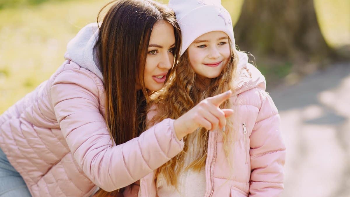Smiling young woman in warm pink jacket leaning towards adorable girl while pointing at something and looking away with curiosity during stroll in city park in sunny spring day