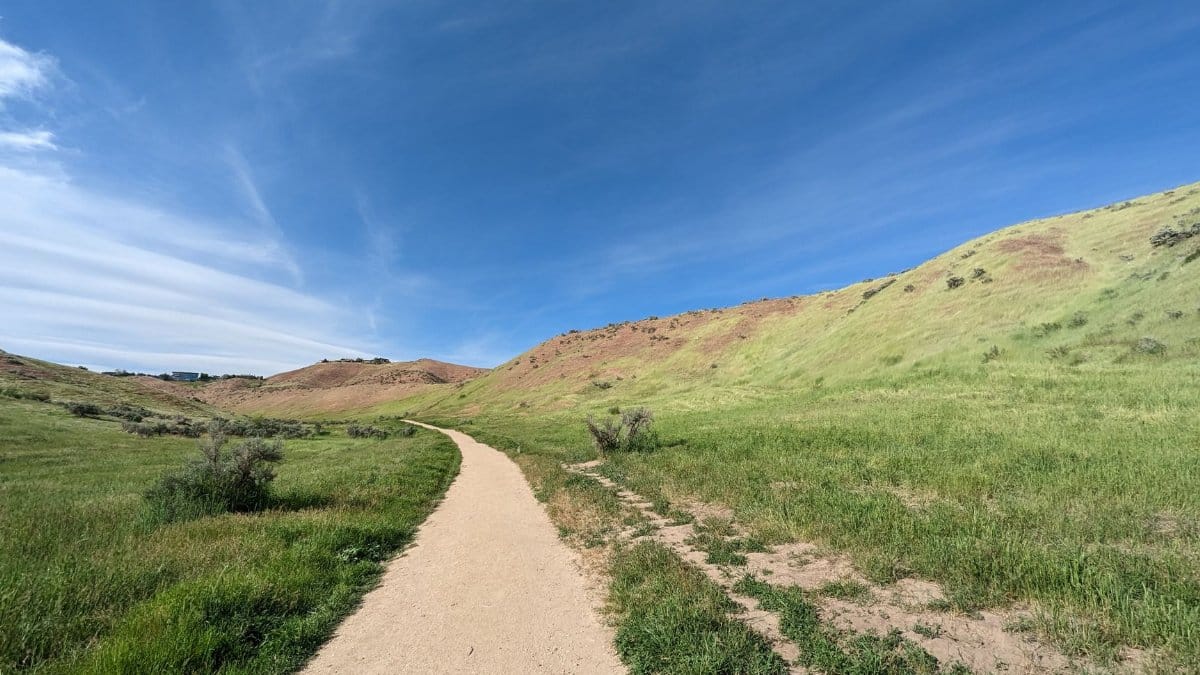 Explore a serene dirt path through the scenic hills and green fields of Boise, Idaho under bright blue skies.