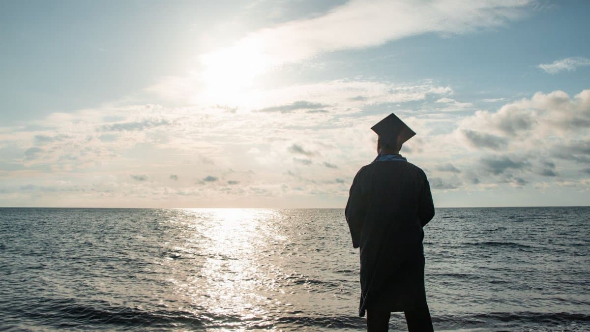 Graduate in cap and gown contemplating the future by the sea at sunset.