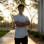 A young man stands confidently on a university campus at sunset, framed by modern architecture and lush trees.