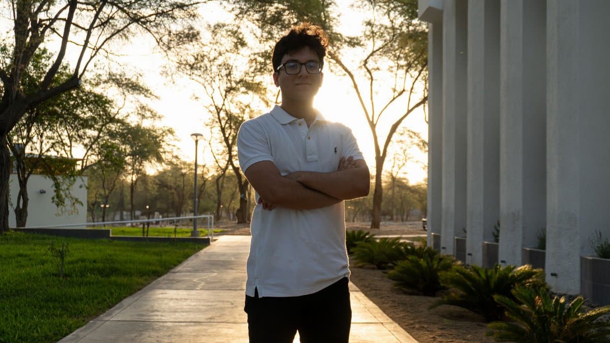 A young man stands confidently on a university campus at sunset, framed by modern architecture and lush trees.