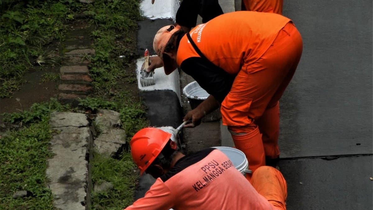 Three workers painting a curb in Jakarta, wearing orange uniforms, in a street maintenance task.