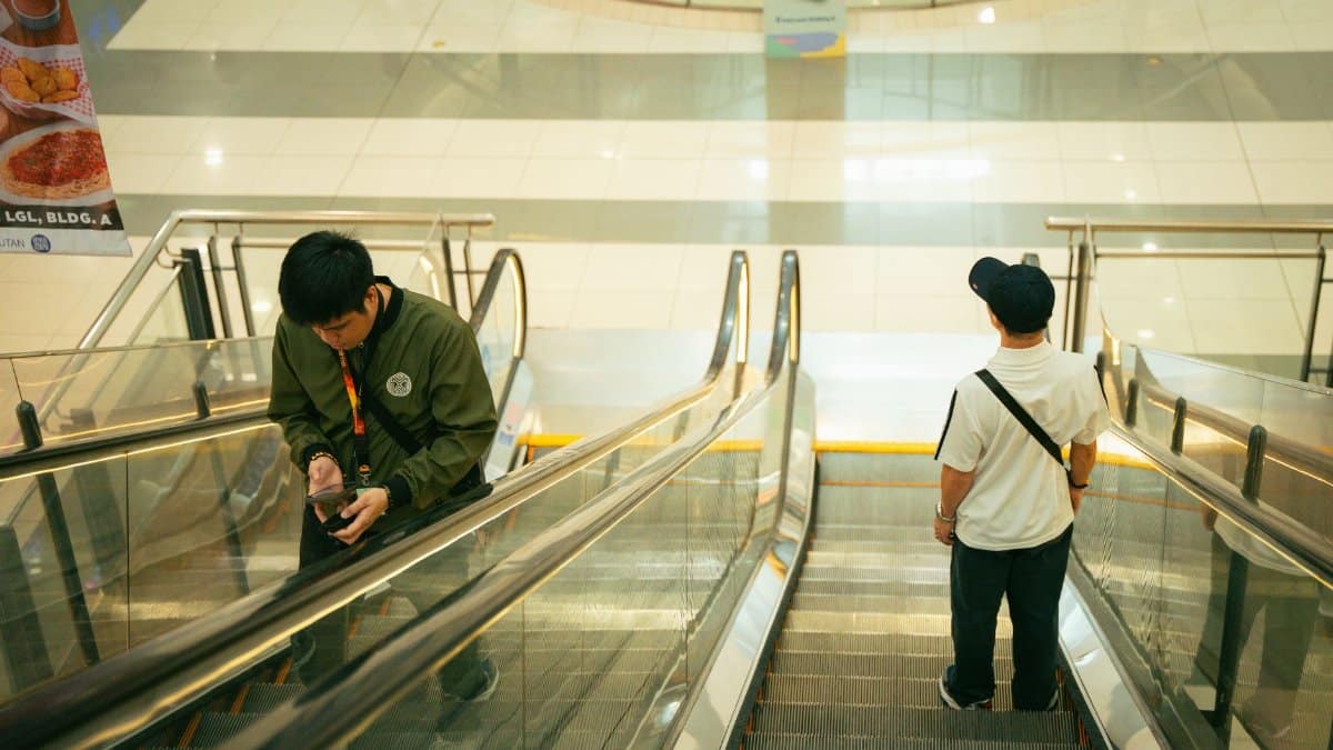 Two men with smartphones on an escalator in a well-lit indoor shopping mall.