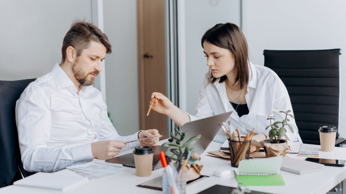 Two colleagues discussing work at a desk with laptops and coffee in a modern office setting.