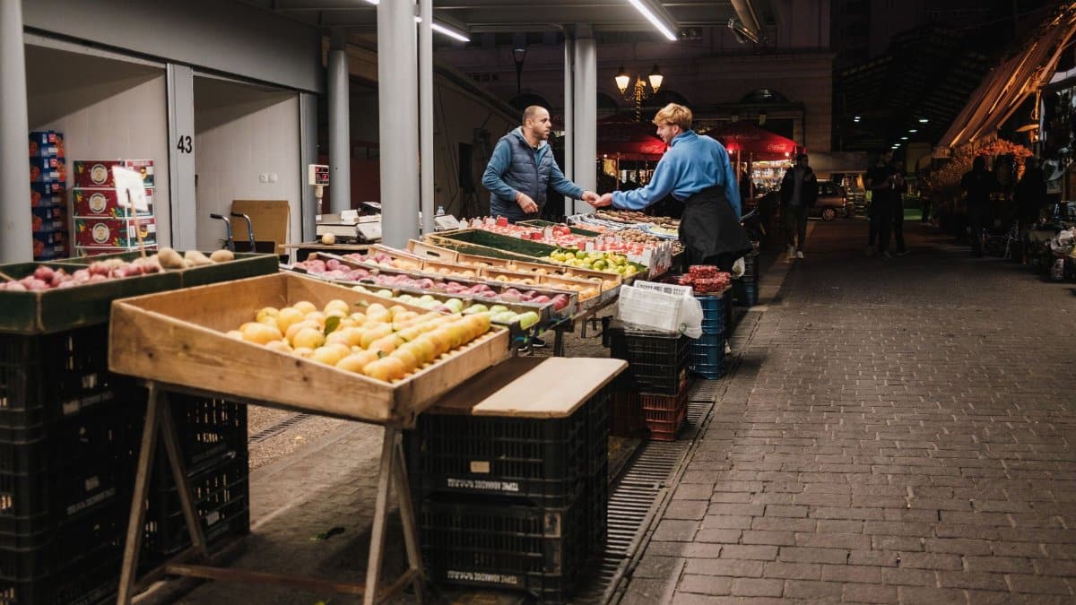 Two people exchanging goods at a well-lit night market in the city, surrounded by fresh produce.