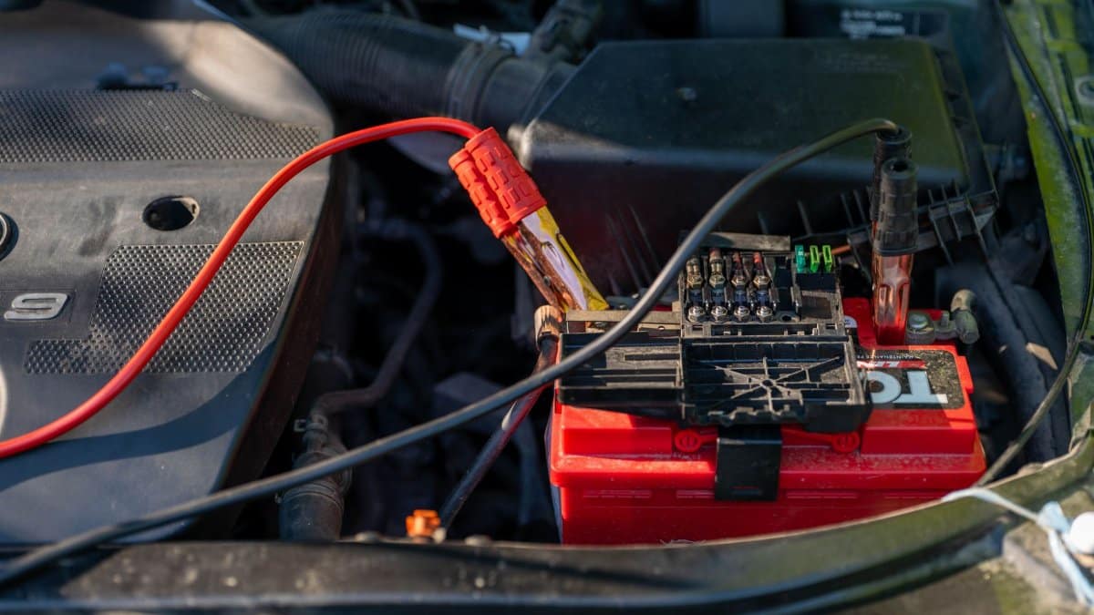 Close-up of a car battery with attached jumper cables in an engine bay.
