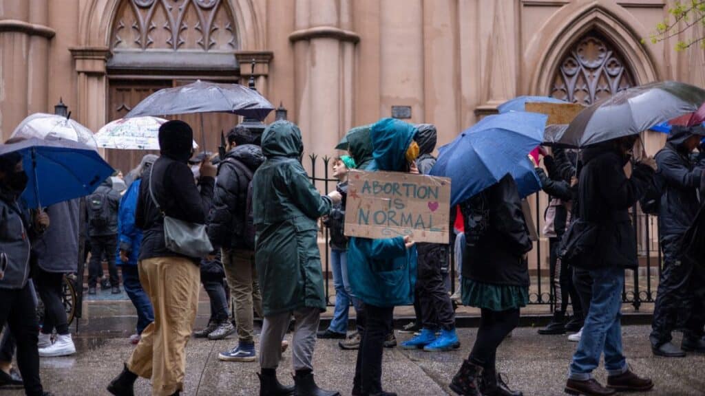 Activists protest for abortion rights in a rainy New York street. Umbrellas and placards visible.