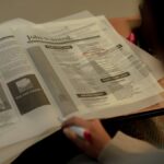 Person reviewing job listings in a newspaper, holding a pink marker for highlighting job ads.