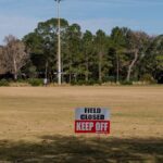 A sports field with a 'Field Closed Keep Off' sign, surrounded by trees and clear skies.