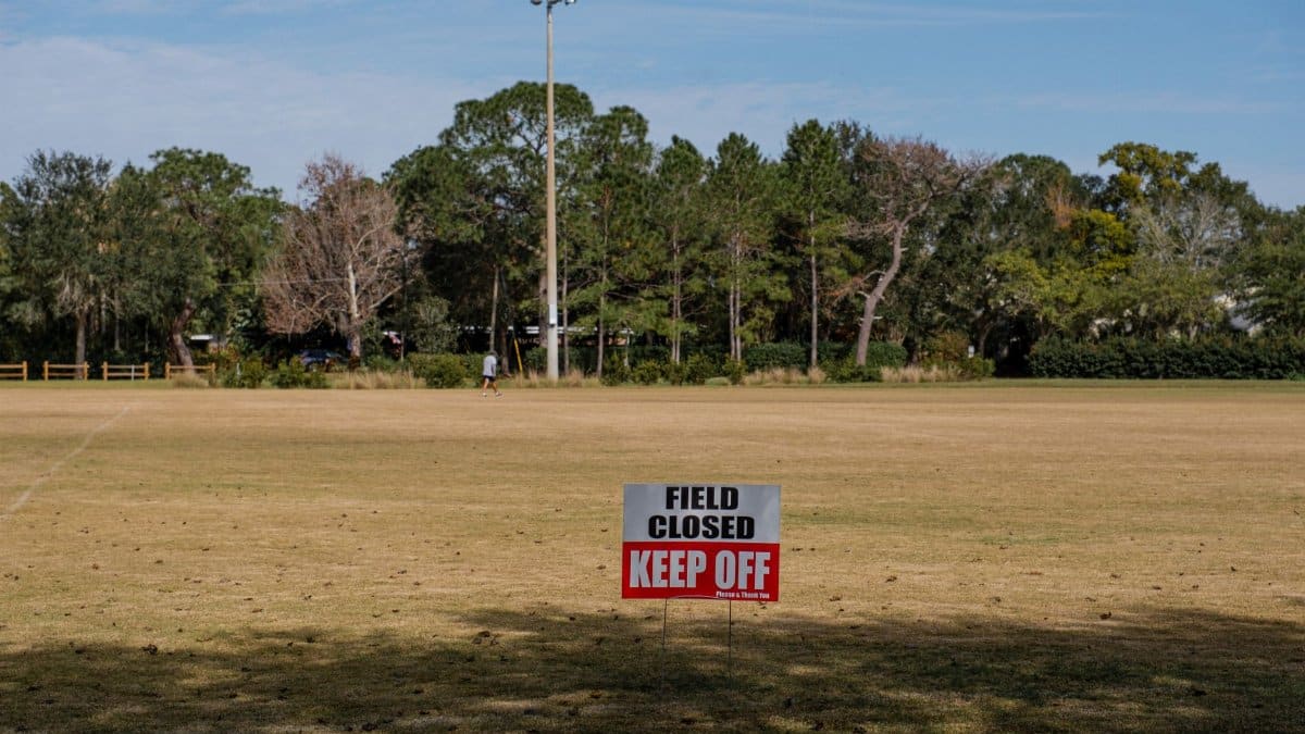 A sports field with a 'Field Closed Keep Off' sign, surrounded by trees and clear skies.