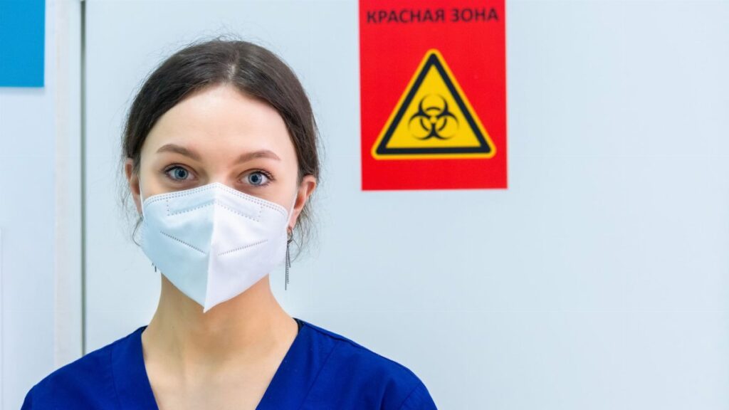 Healthcare professional in protective gear standing near a biohazard warning sign.