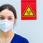 Healthcare professional in protective gear standing near a biohazard warning sign.