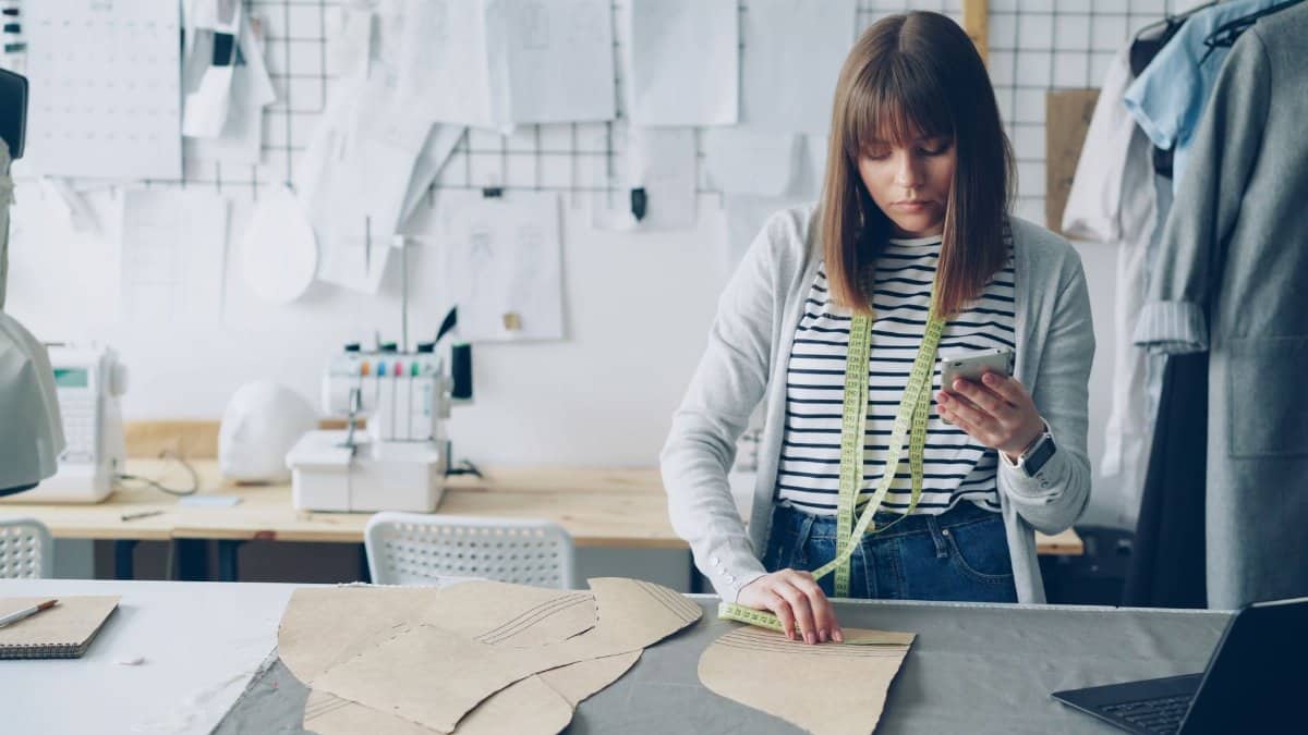 Young female fashion designer using mobile and measuring fabric in a creative workshop.