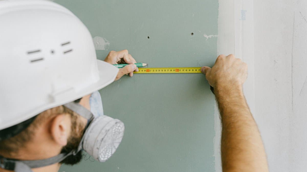 A construction worker precisely measuring drywall, wearing a helmet and mask for safety.