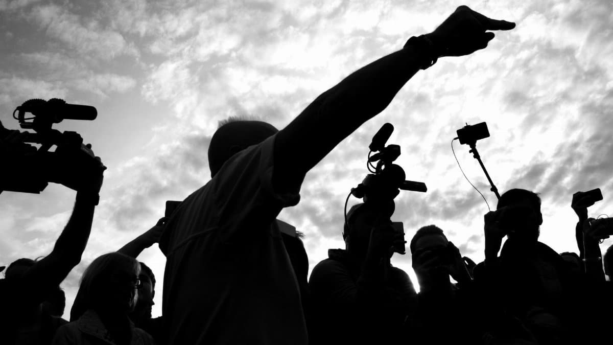 A dramatic silhouette of a media crew with cameras capturing a pointing individual against a cloudy sky.
