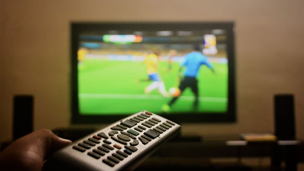 Close-up of a hand holding a remote control in front of a TV showing a football match.