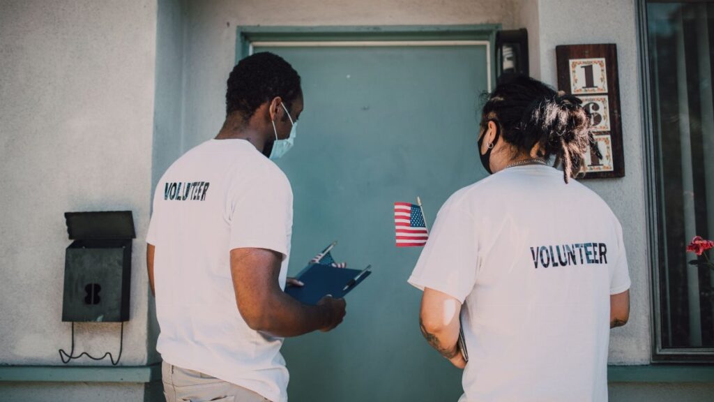 Two volunteers wear masks and visit community homes, engaging in outreach activities.