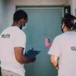 Two volunteers wear masks and visit community homes, engaging in outreach activities.