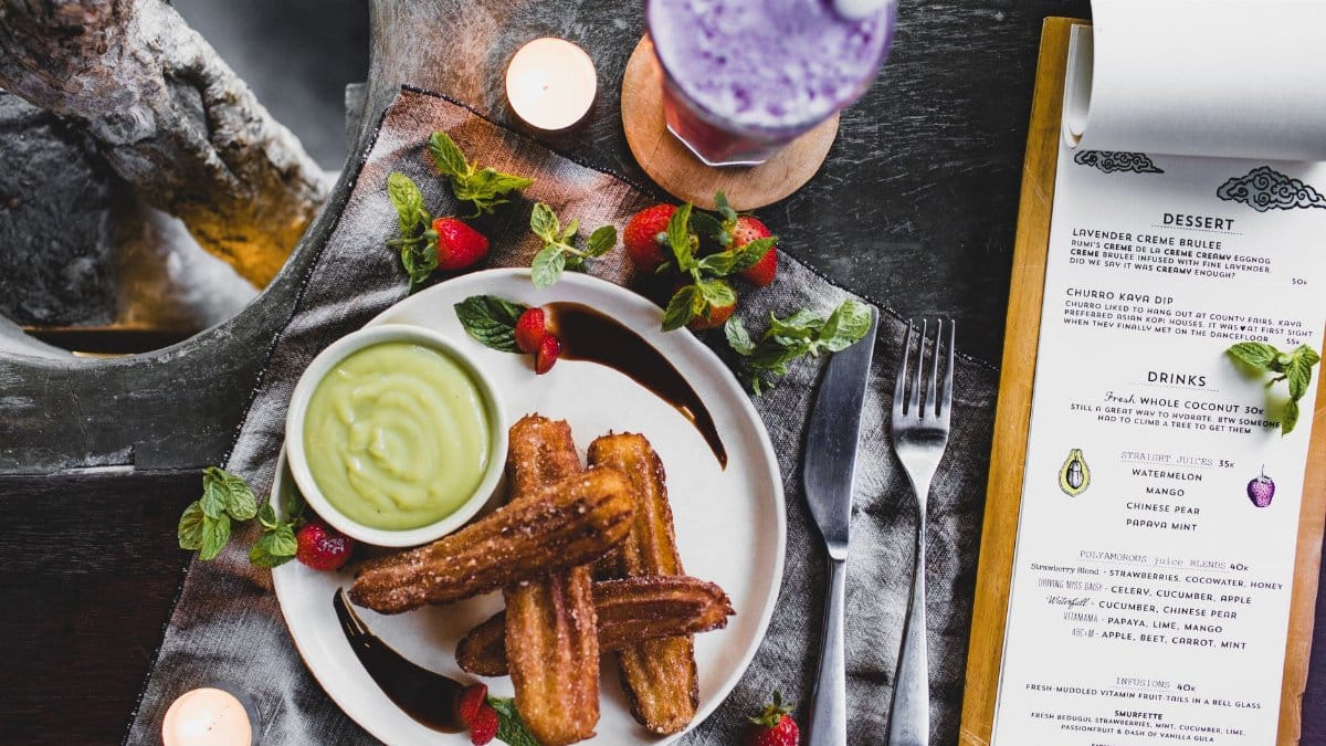 From above of delicious Spanish churros with green dip sauce garnished with fresh strawberries and melted chocolate near glass of blueberry milkshake and menu in restaurant
