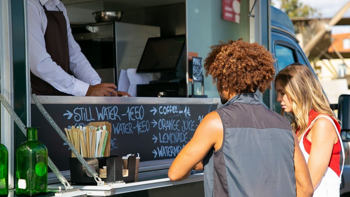 Multiethnic couple standing together near food truck with seller and selecting dishes from menu in sunny day